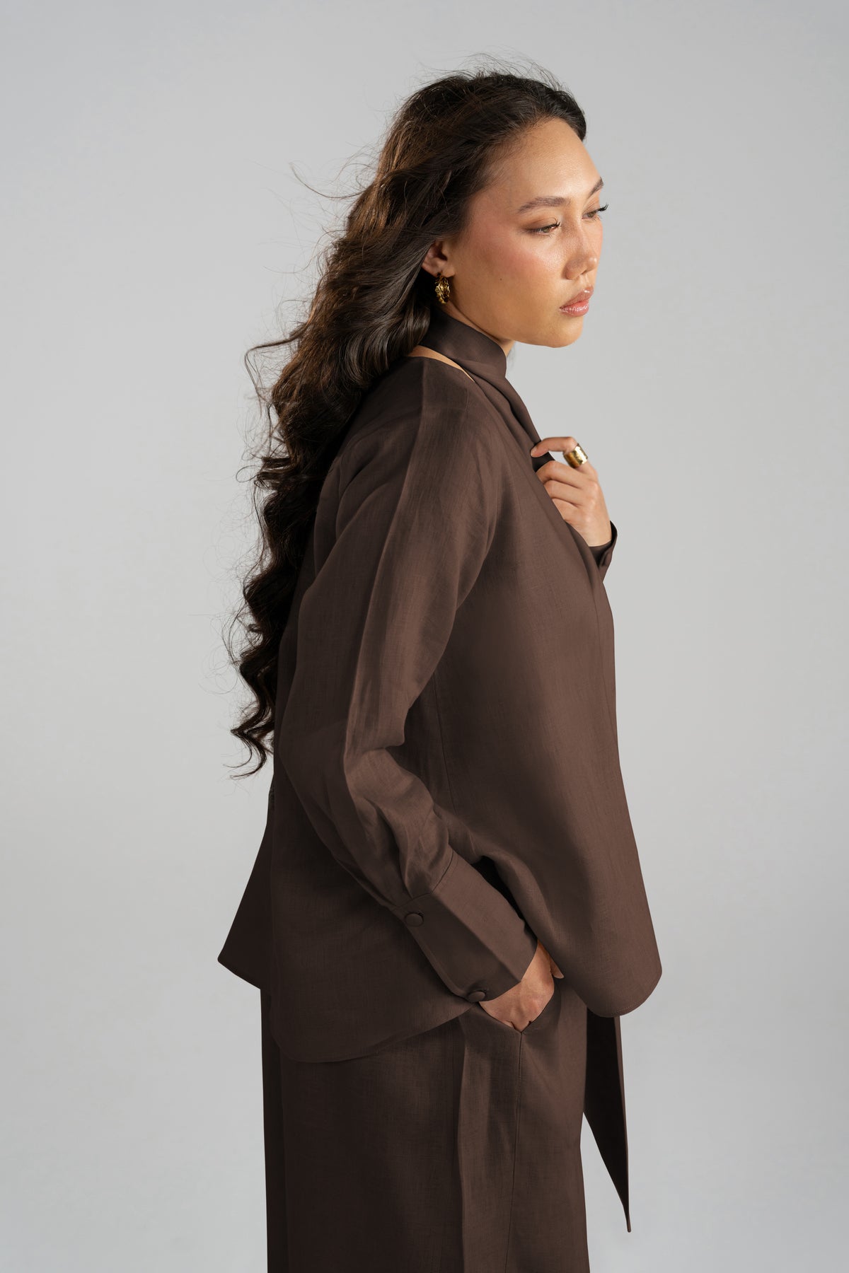 A woman in a side profile wearing a dark brown linen high-neck long-sleeve blouse and matching trousers